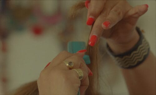 A woman's hands with orange nail polish, teasing her hair with a comb.